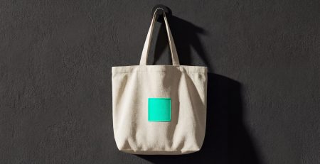 A premium minimalist canvas tote bag with a single neon-teal square emblem hanging on a dark textured wall, illustrating the CPG brand community tote-bag test.