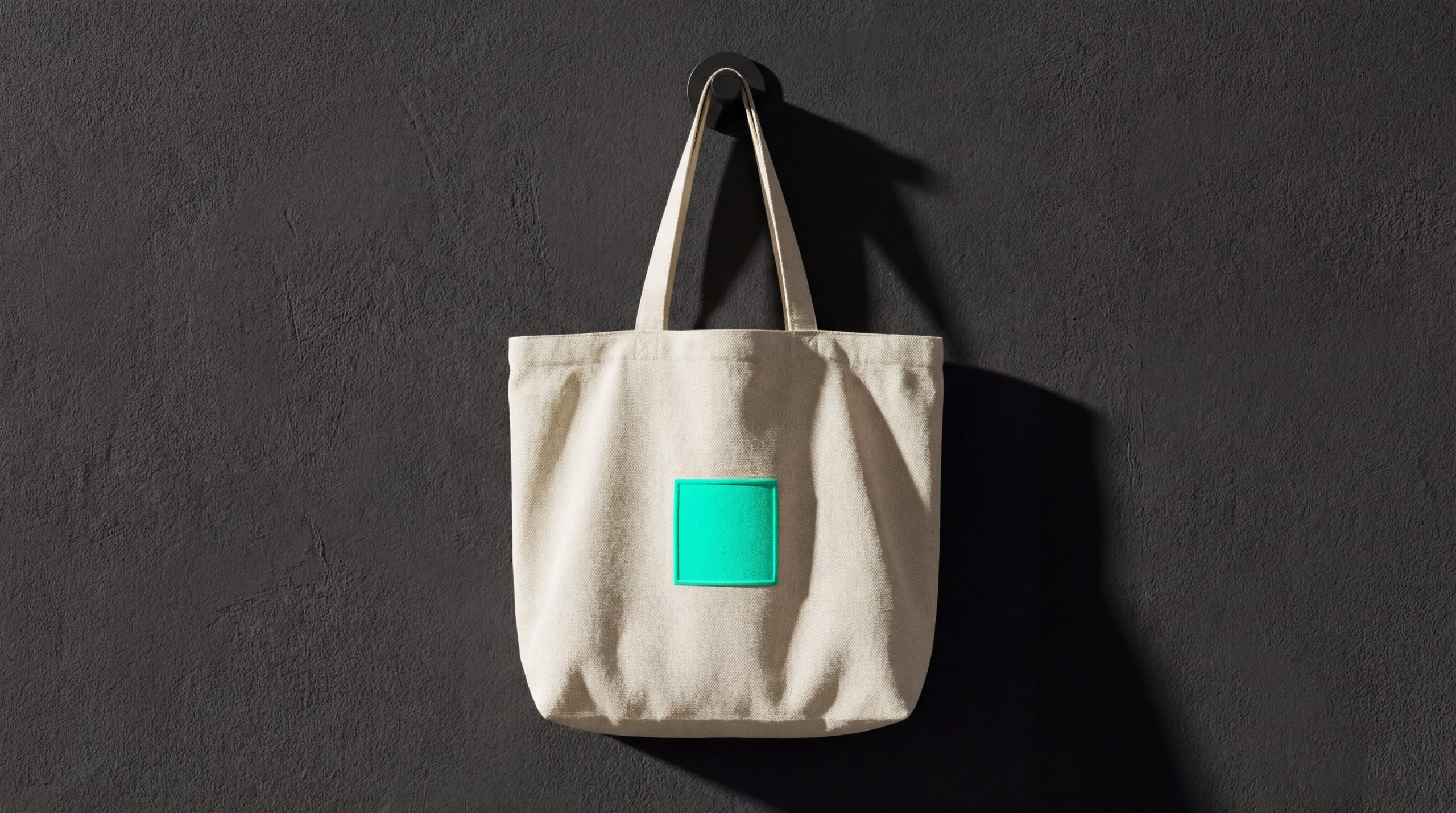 A premium minimalist canvas tote bag with a single neon-teal square emblem hanging on a dark textured wall, illustrating the CPG brand community tote-bag test.